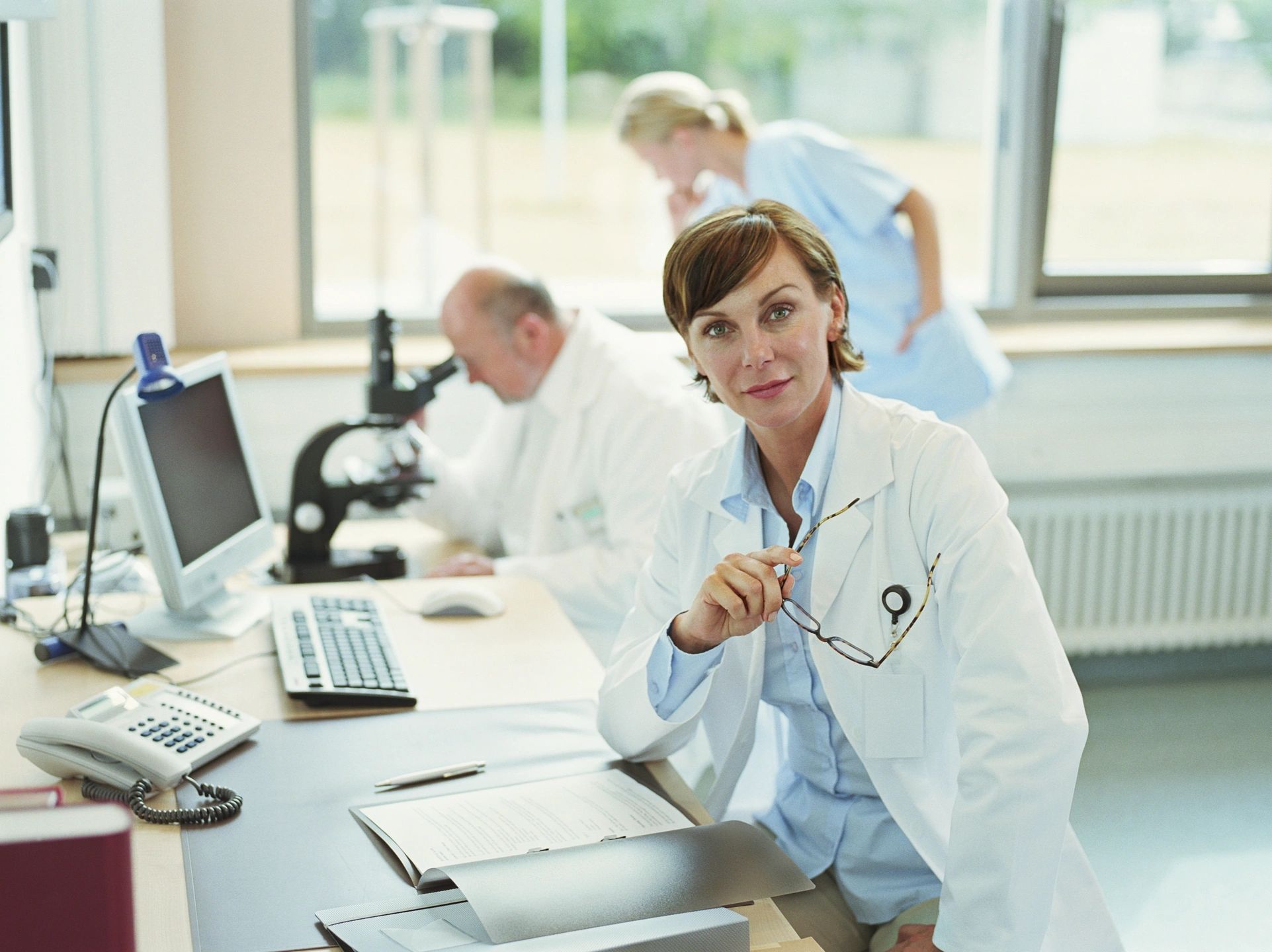 Female doctor with file in front of them with two other doctors at a microscope in the background.